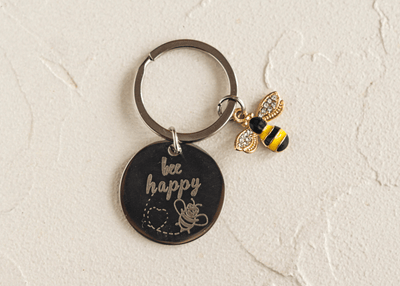 "Schlüsselanhänger mit 'bee happy' Gravur und funkelnder Bienenfigur auf cremefarbenem Hintergrund, symbolisiert Freude und Naturverbundenheit."
